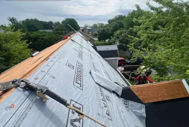 Roof under repair with silver underlayment and workers on a steep rooftop beside trees and houses