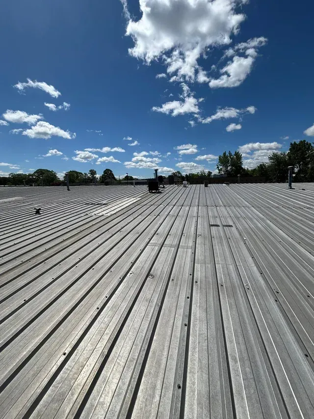Rows of gray metal roofing under a bright blue sky with scattered clouds, stretching toward a distant treeline.
