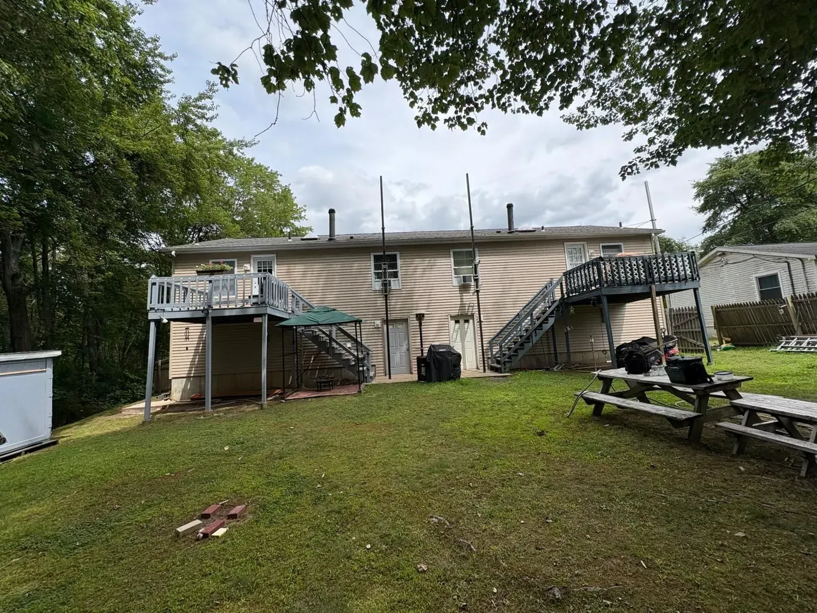Backyard with a small beige house, dual staircases, a grassy lawn, and picnic tables under trees.