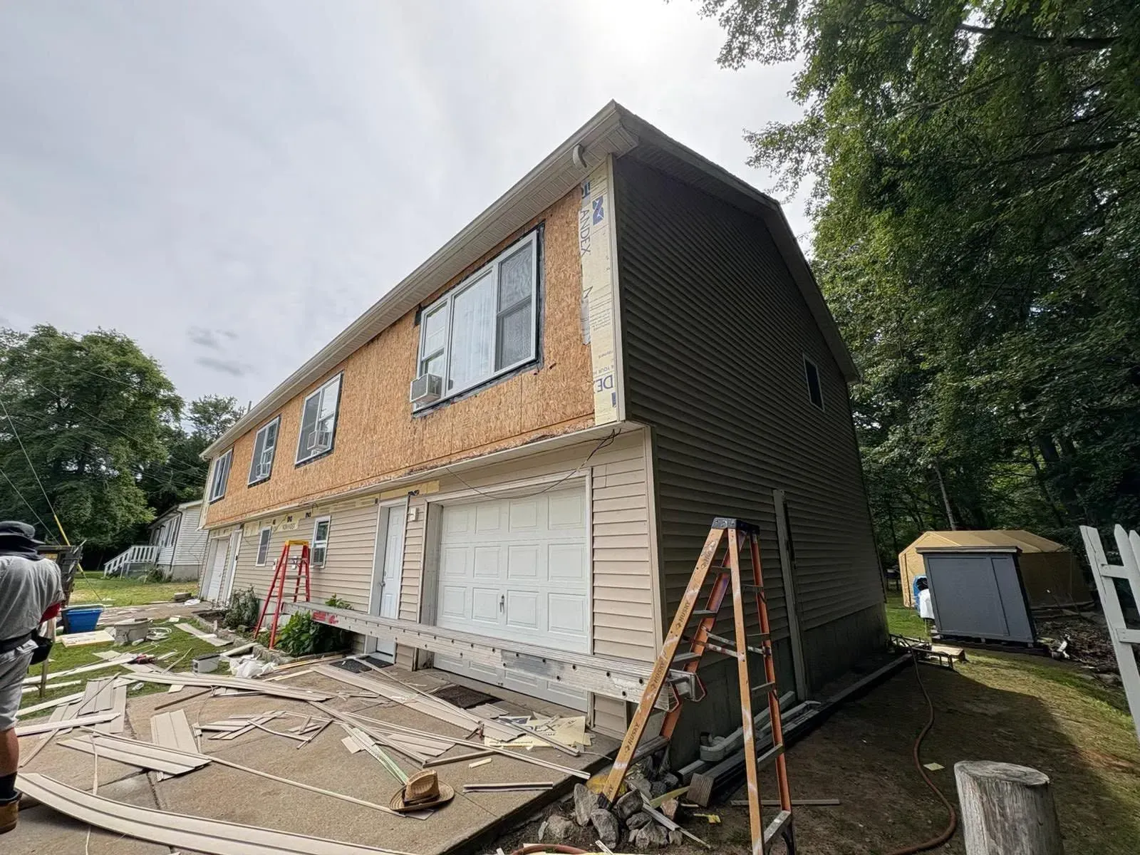 Two-story house under renovation with exposed siding, garage doors, and construction debris in the yard