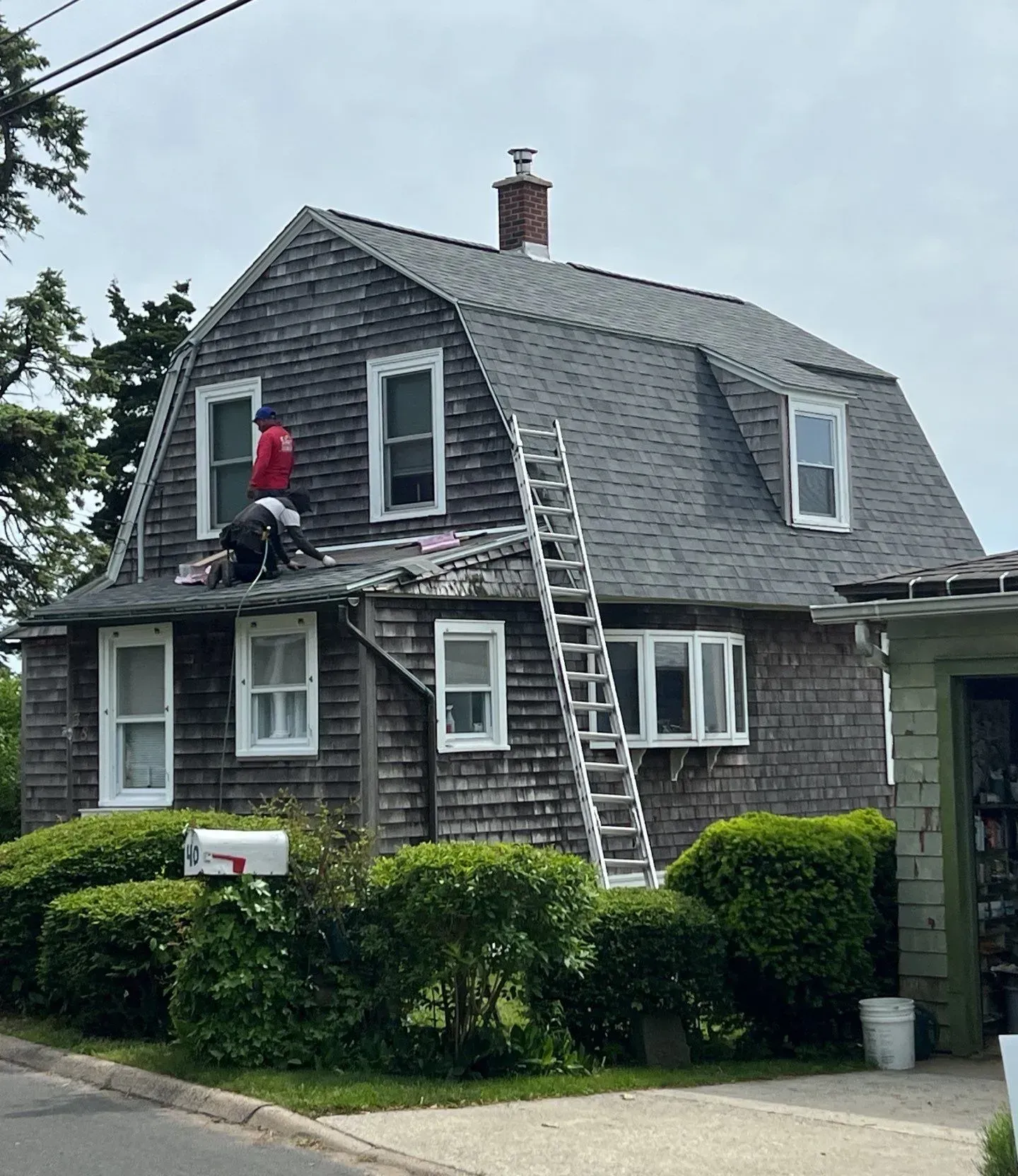 Workers on a roof repairing a gray house, with ladders and bushes in front
