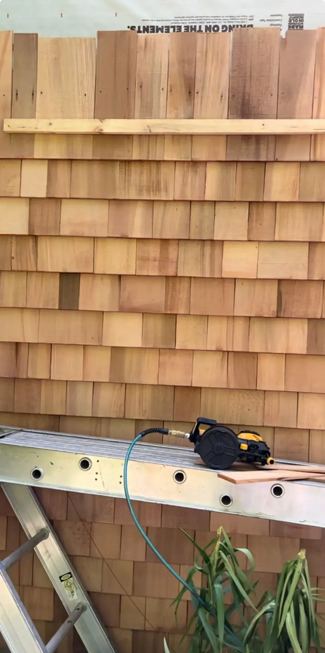 Wooden shingle wall under construction with a power drill on a workbench and plants below