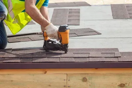 Roofer uses a nail gun to install asphalt shingles on a wooden roof; wearing gloves and safety vest.