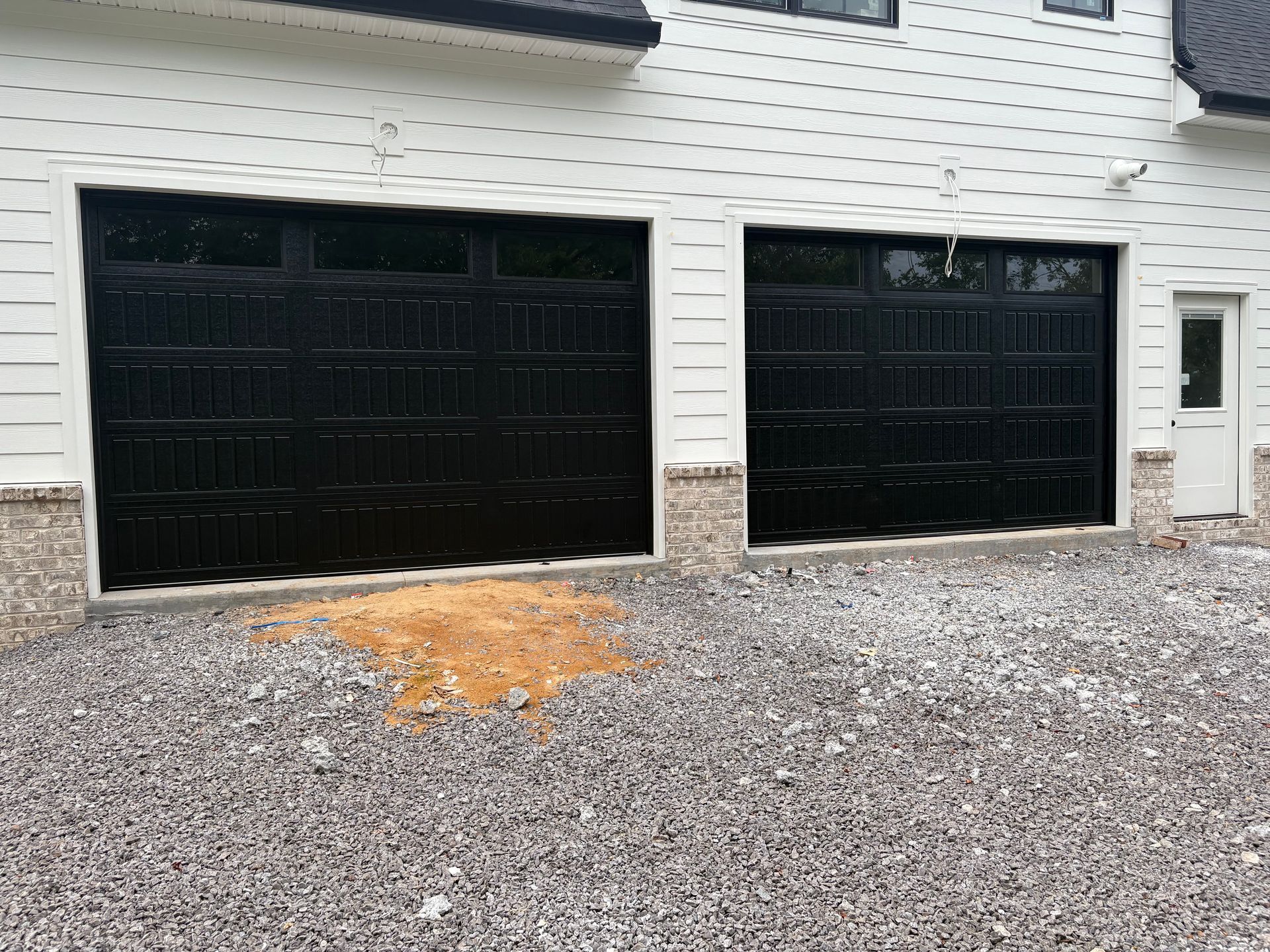 Black garage doors on a white building, gravel ground. A small door is visible to the right.