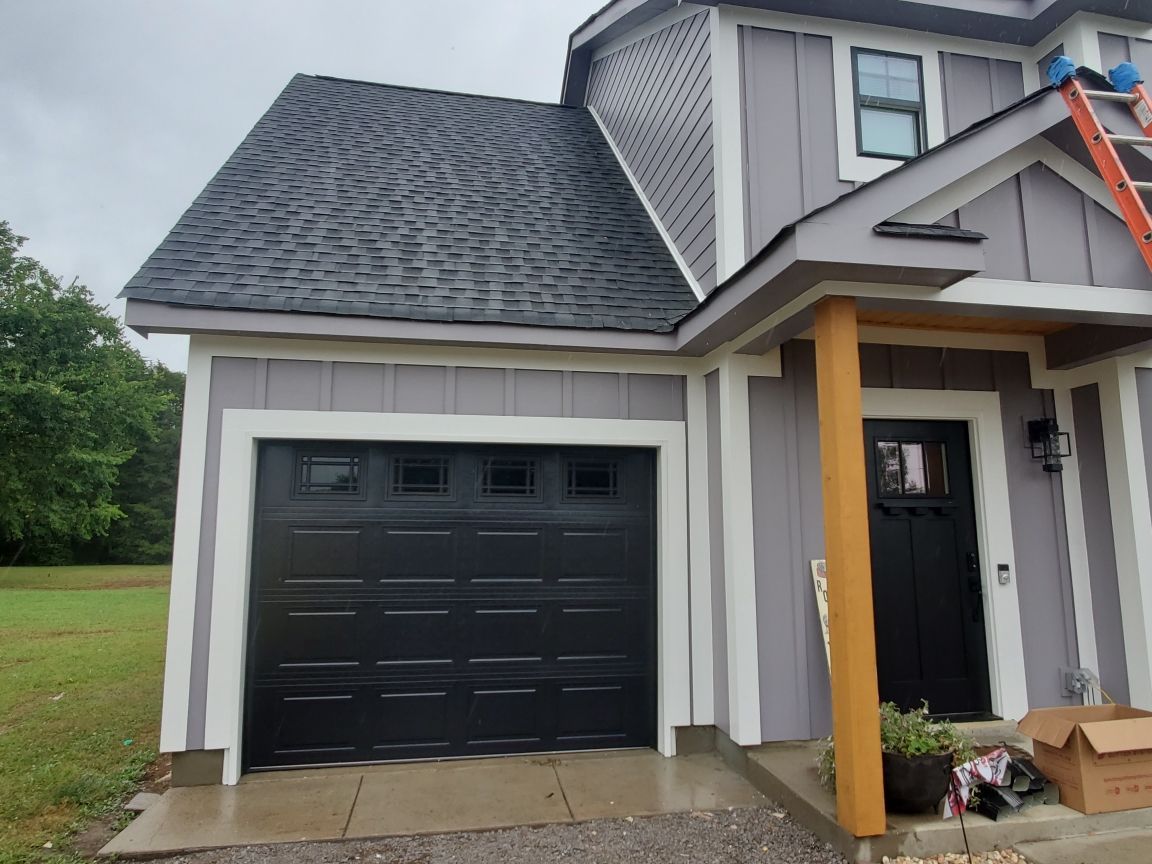 Gray house with black garage door, front door, and roof. Wooden beams and green yard.