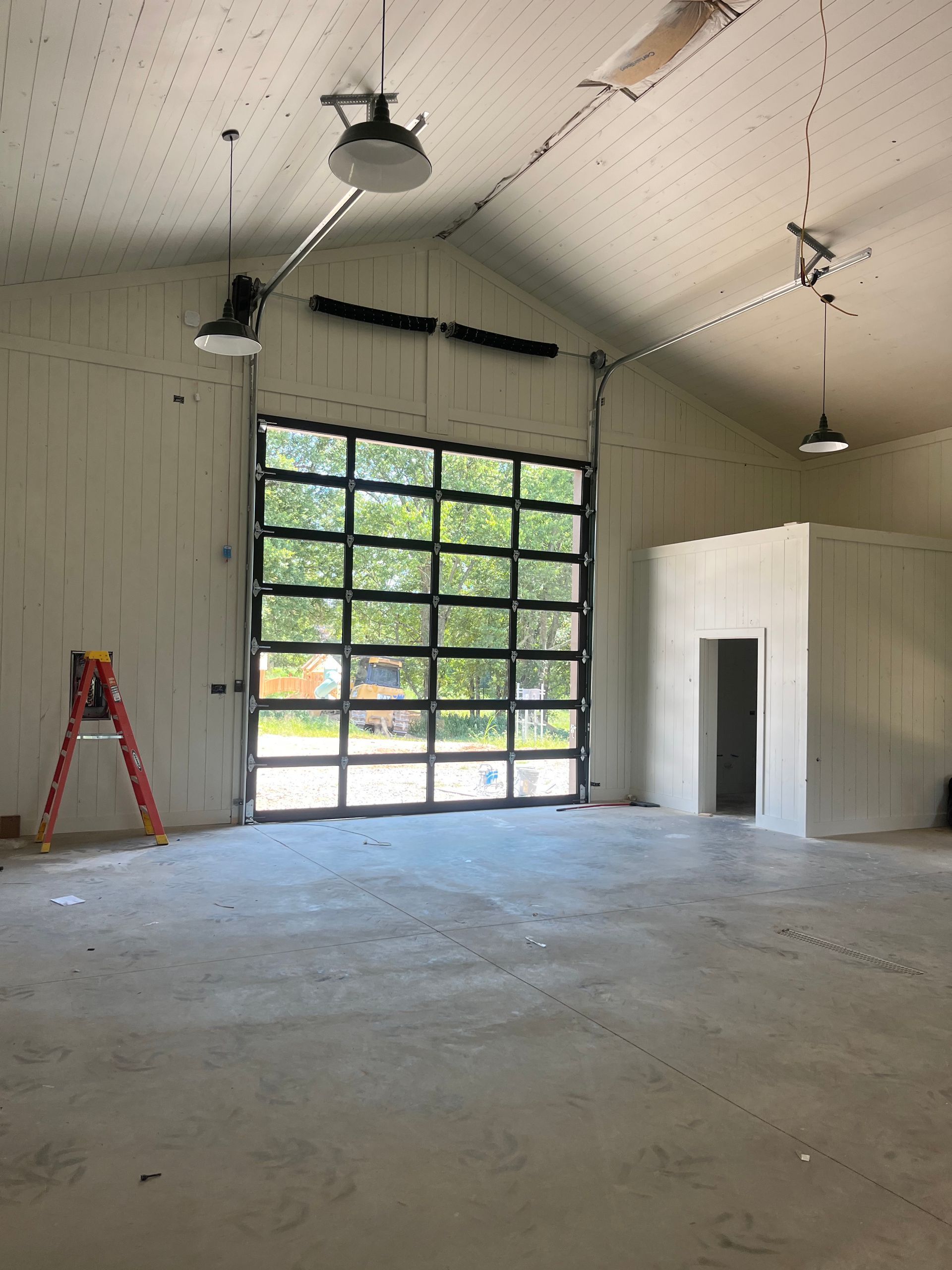 Large empty building interior with glass garage door, ladder, and concrete floor.