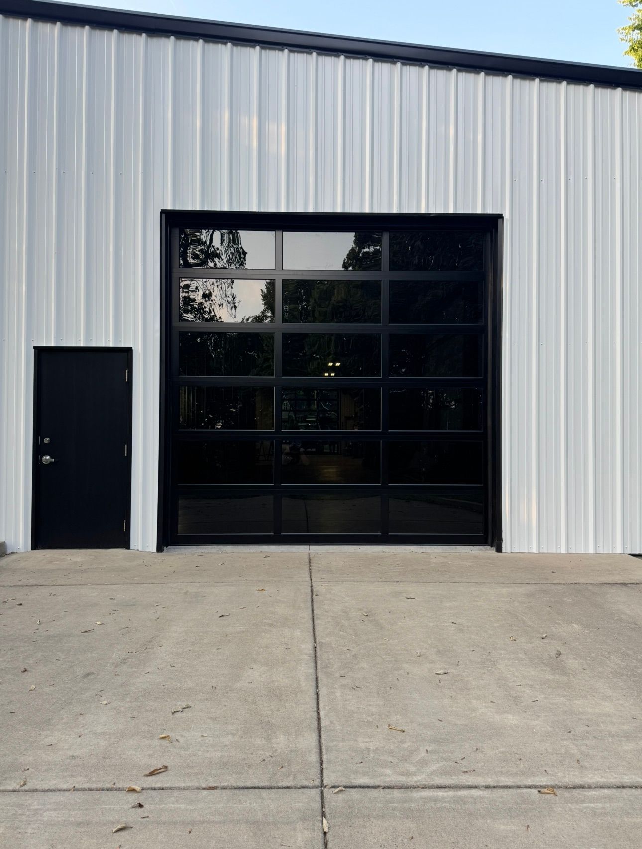White metal building with a black-framed glass garage door and a black side door, set on a concrete pad.