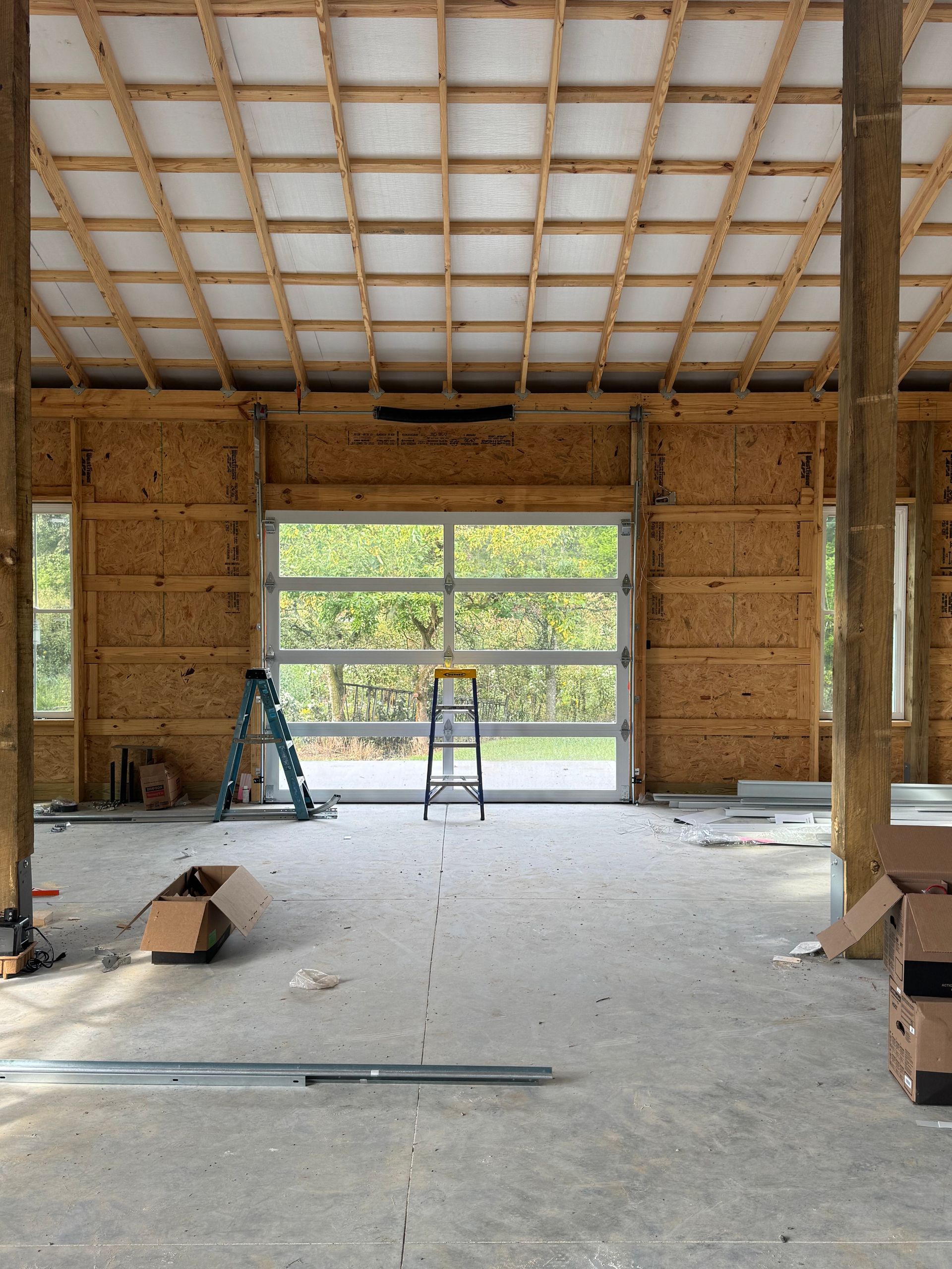 Interior of a building under construction. A large glass garage door faces the outside. Exposed wooden beams and rafters.