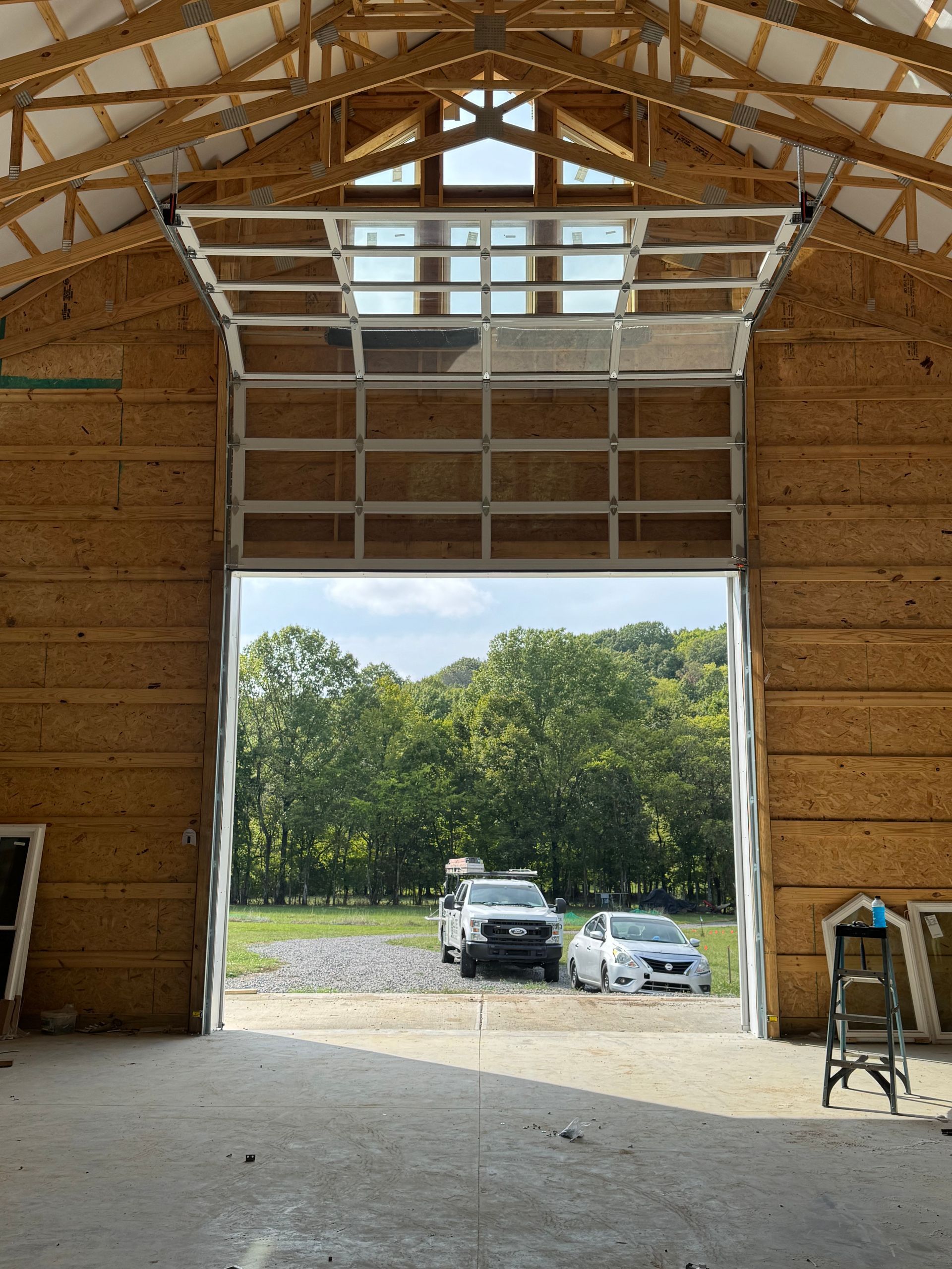 Open garage door in wood-framed building, revealing cars on gravel drive and green trees.