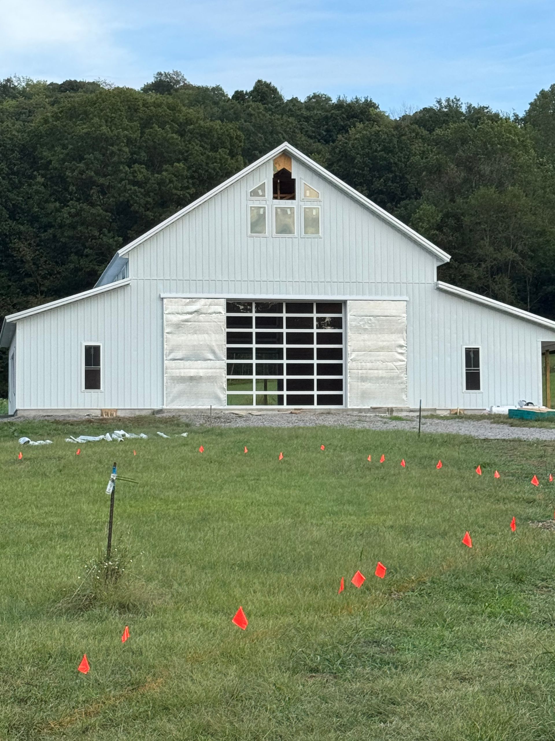 White barn with large glass door, under construction, set in a grassy field with trees in the background.
