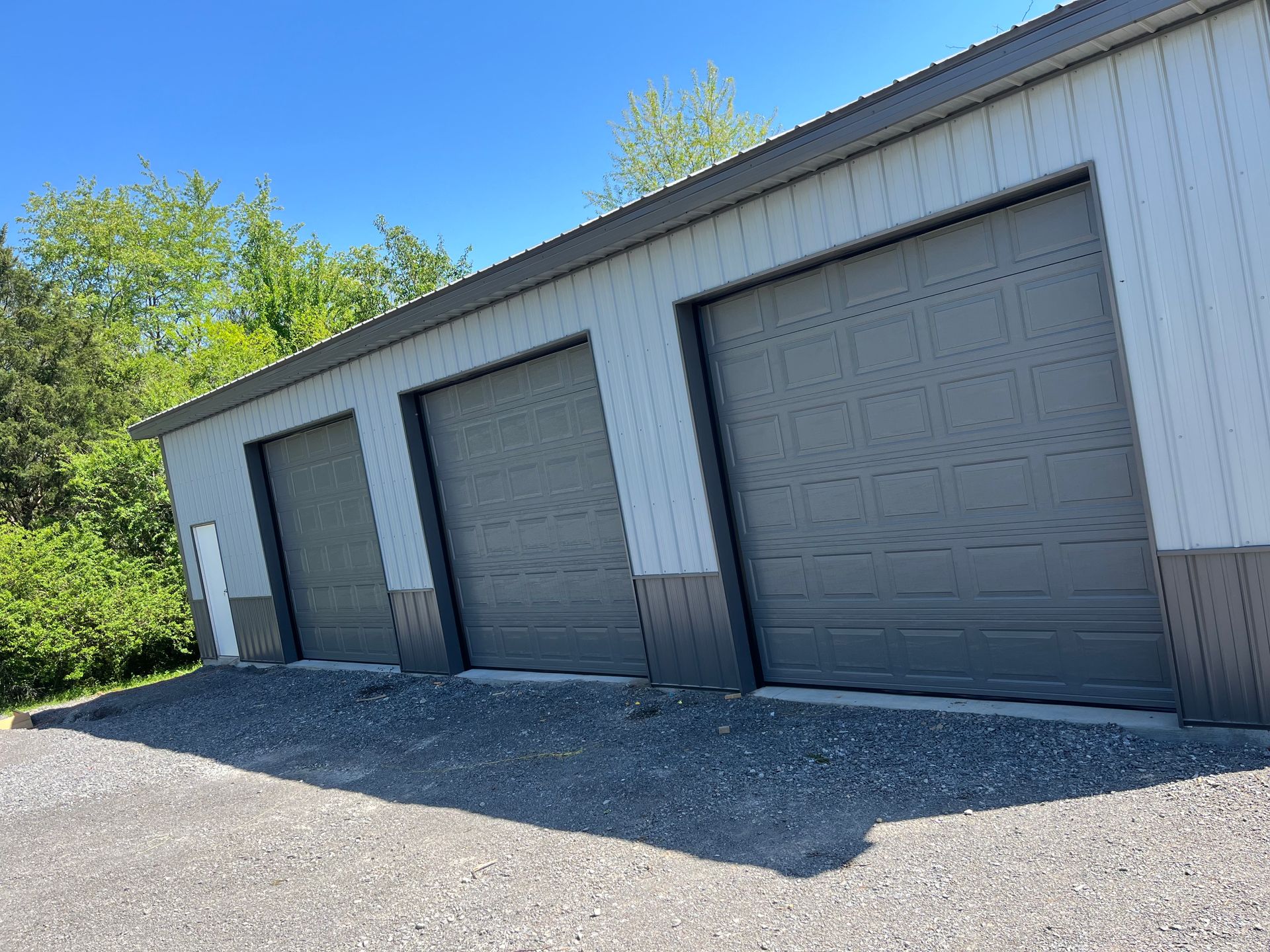 Three gray garage doors on a metal building with gravel driveway under a blue sky.