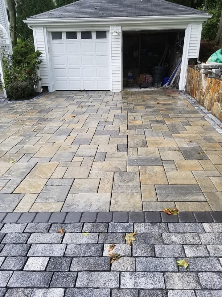 A paved driveway with a garage in the background. Driveway made with rectangular blocks of tan, gray, and black.