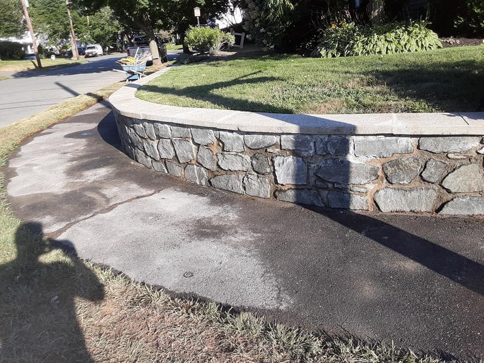 Curved stone retaining wall bordering a paved sidewalk and a grassy area. Person's shadow visible.
