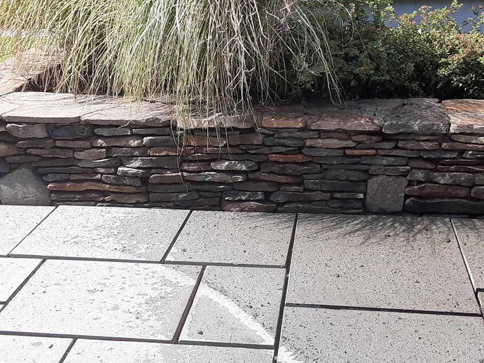 Stone retaining wall with plants above, next to stone patio.