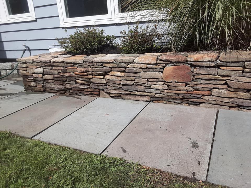 Stone retaining wall with plants, bordering a gray patio and green grass. Blue house in background.