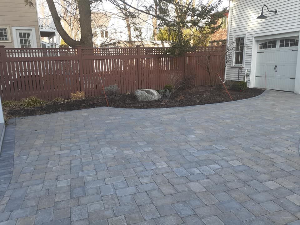 Brick driveway leading to a garage with a wooden fence and garden bed.