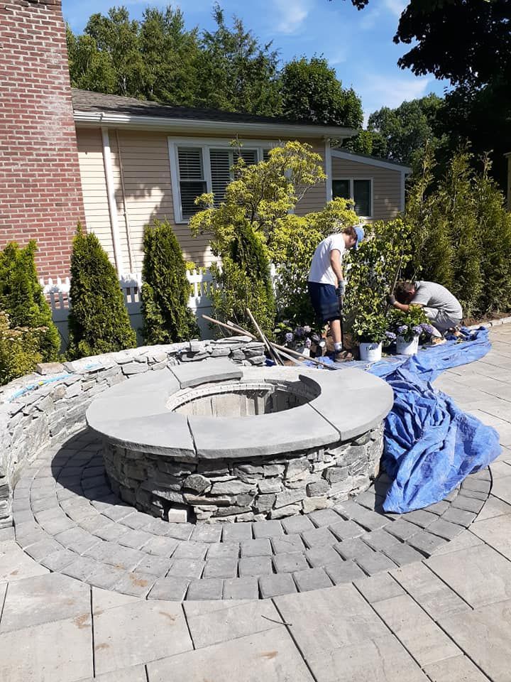 Circular stone fire pit in a brick patio, with two people working nearby a house.