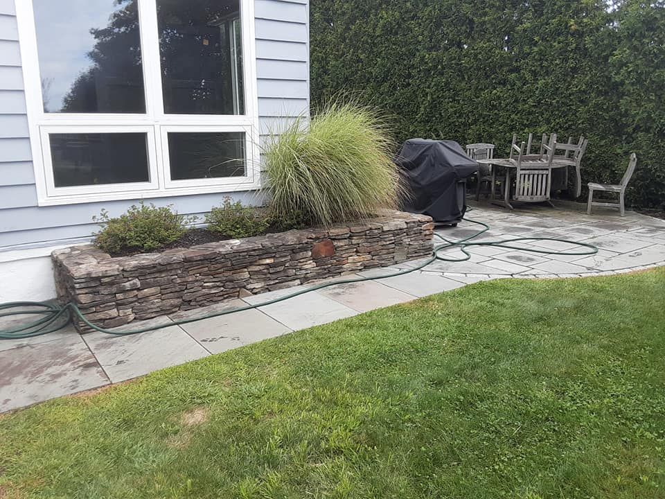 Stone planter with plants next to a house with a window, patio with furniture, and a grassy yard.