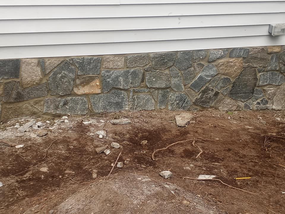 Stone foundation below white siding; dirt and debris in foreground.