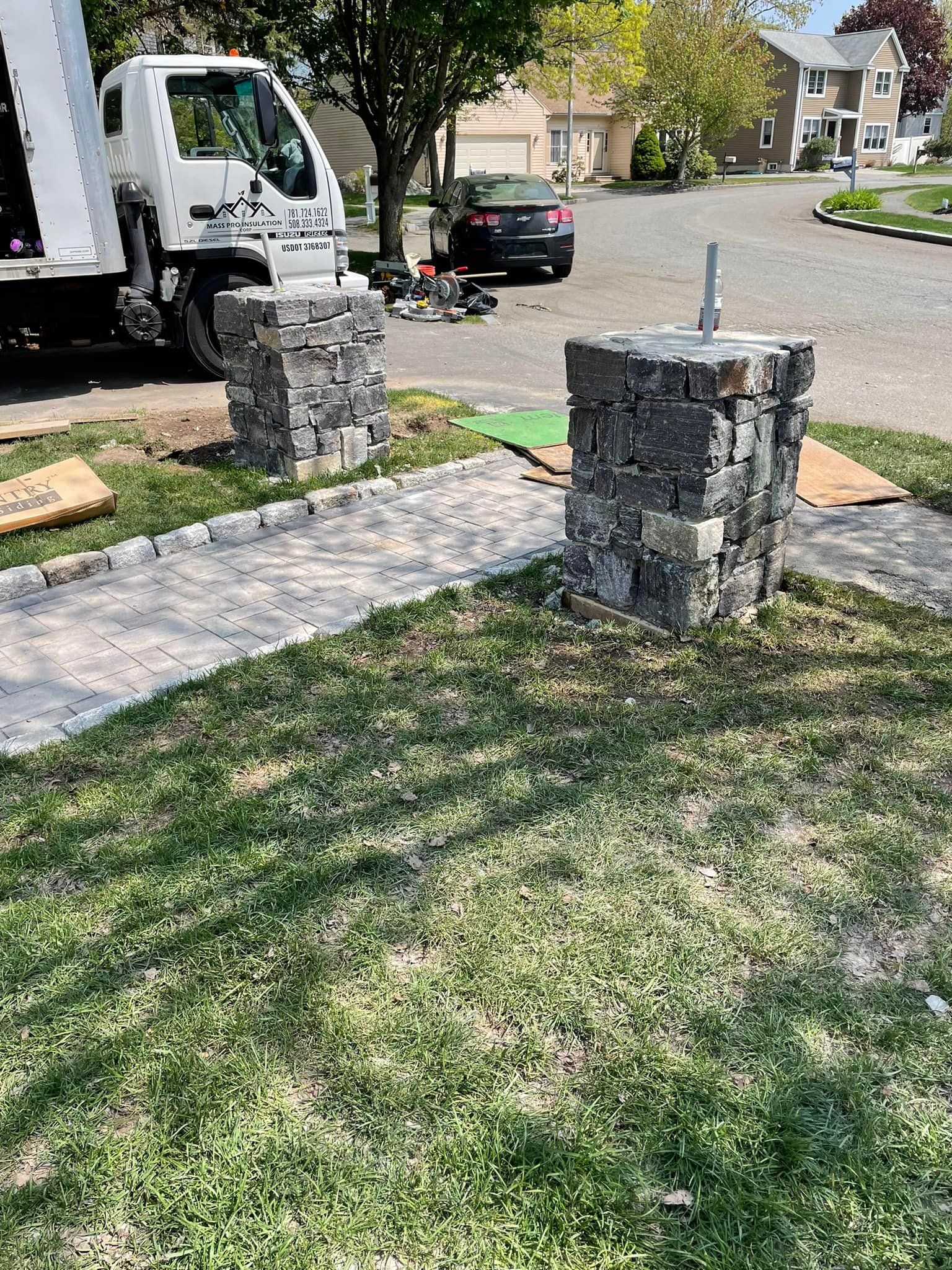 Stone pillars with pathway under construction, beside a lawn and street with parked cars.