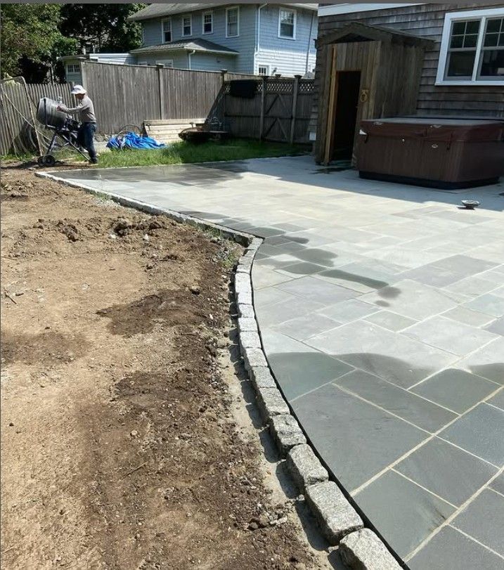 Newly constructed paved patio with stone border, adjacent to dirt, fence, and a house.
