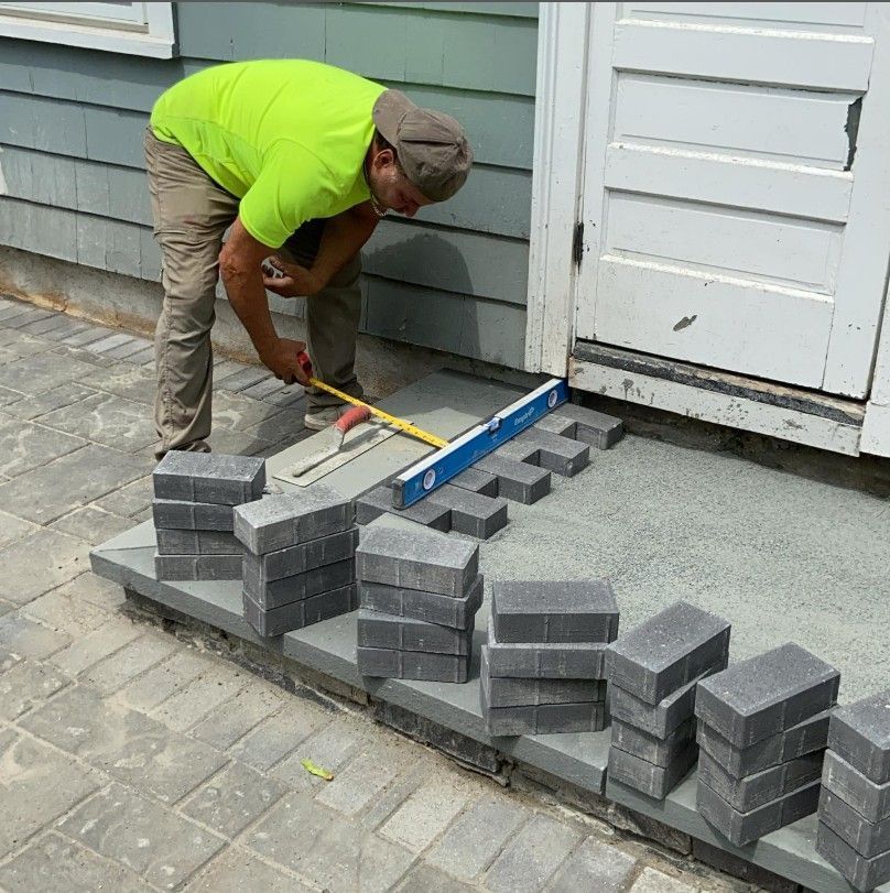 Person laying pavers at a doorway. Gray blocks, blue level, yellow shirt, building with blue siding.