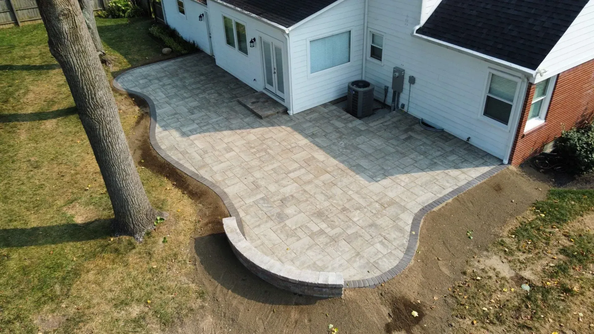Back patio with light stone pavers beside a white house and curved retaining wall