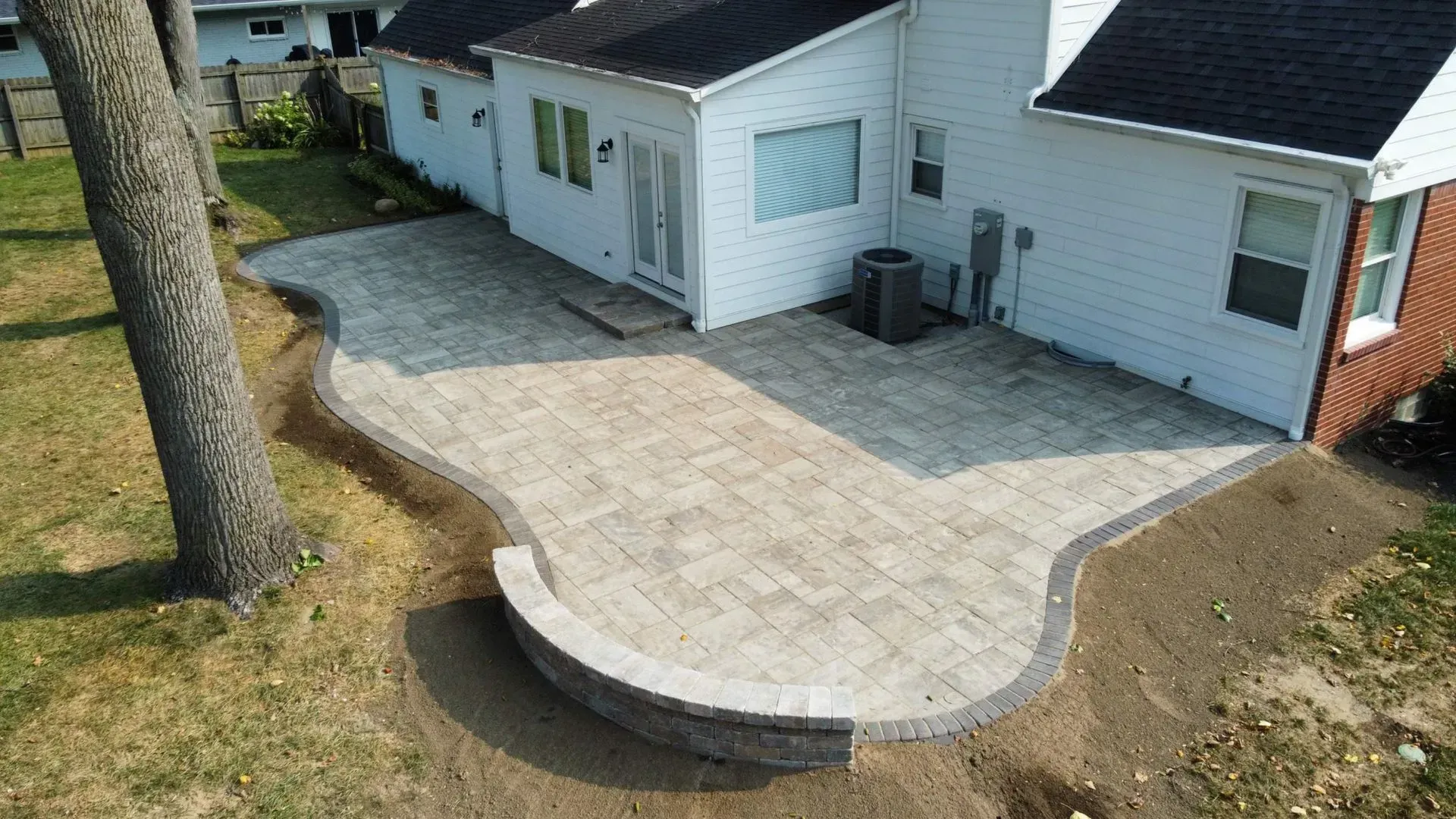 Curved stone patio beside a white house and garage, with a tree and exposed soil edging