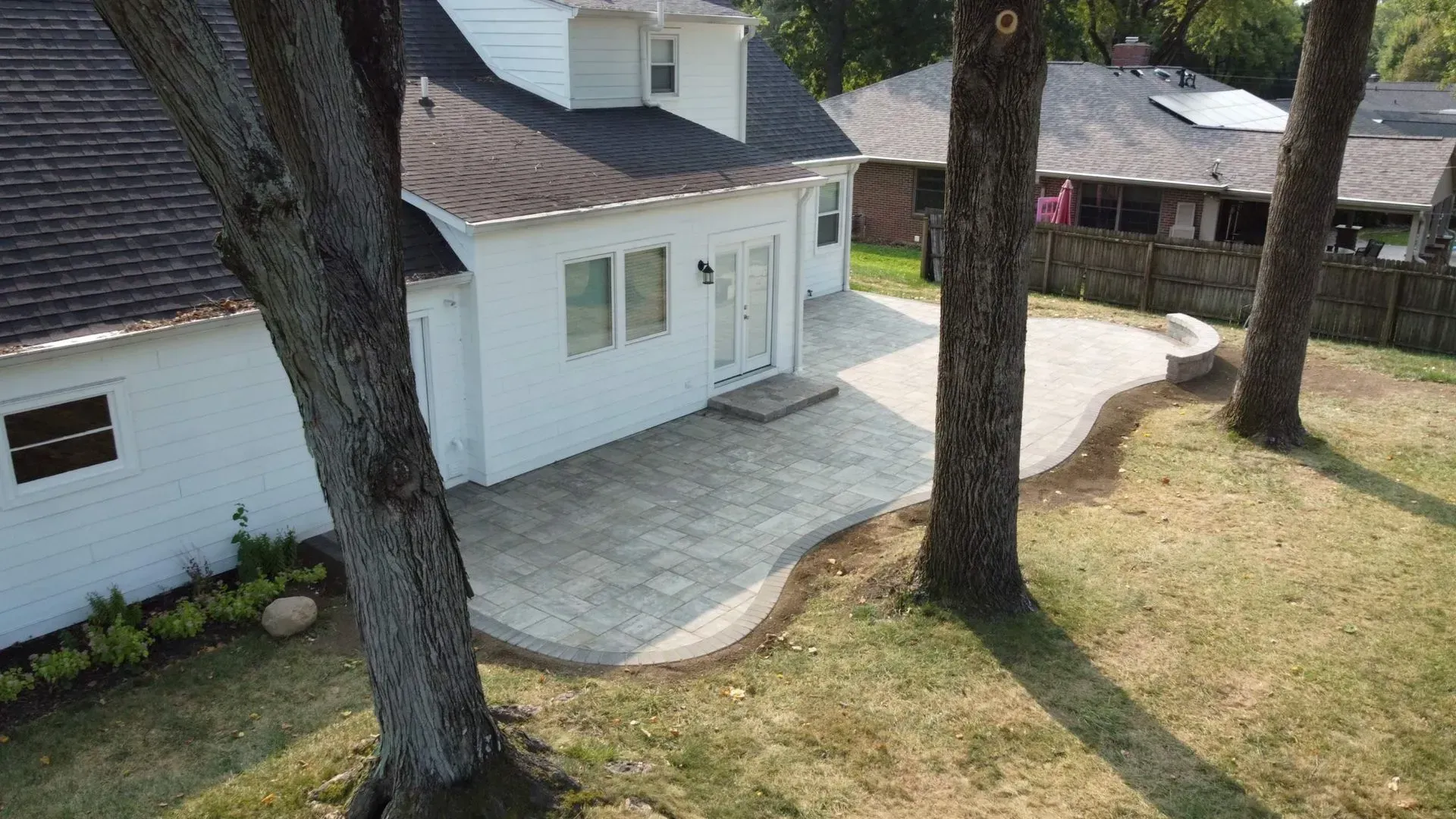 White house with a curved stone patio and large trees in a sloped backyard.