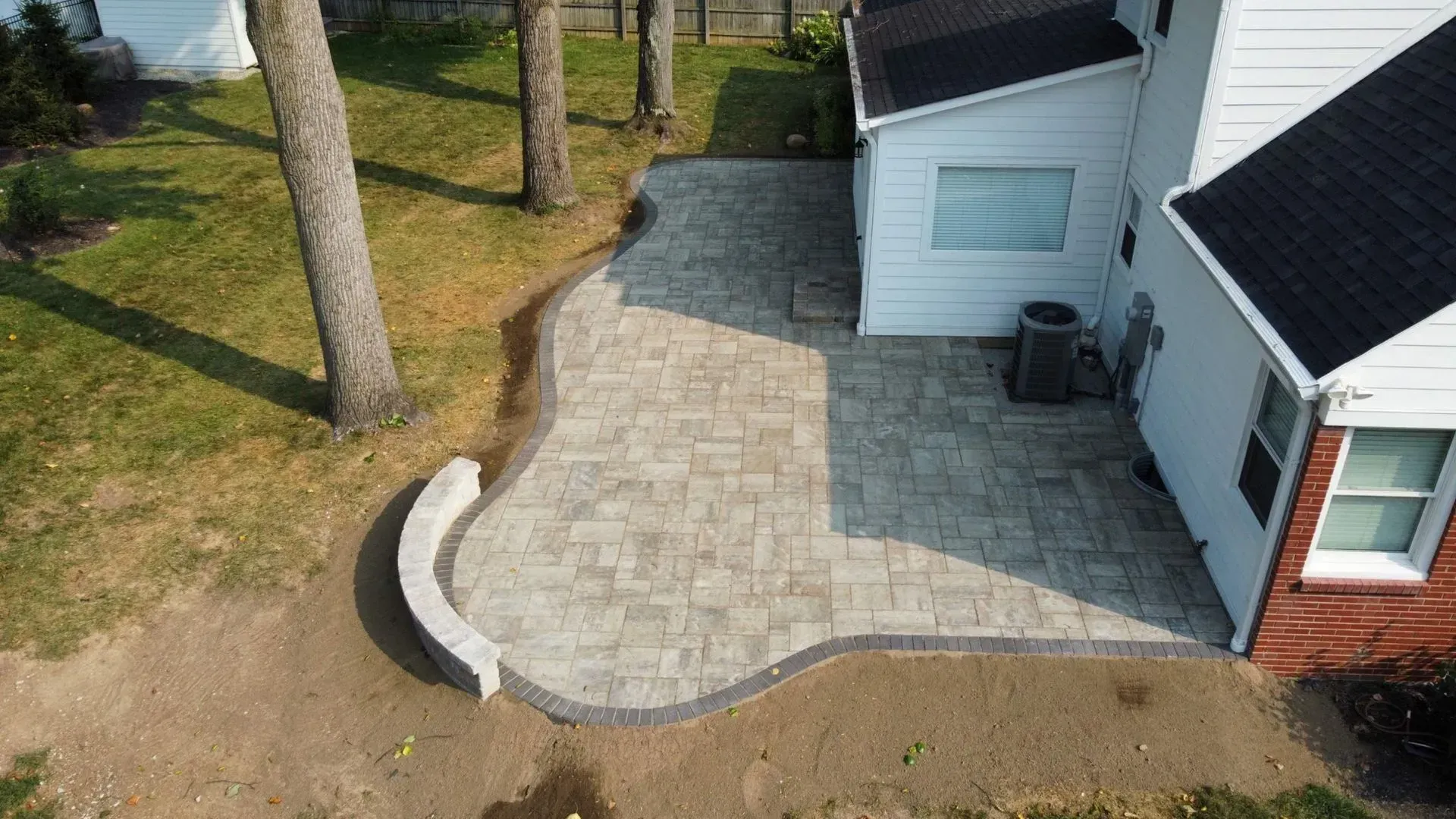 New gray paver patio beside a white house, with a curved concrete bench and a small window well.