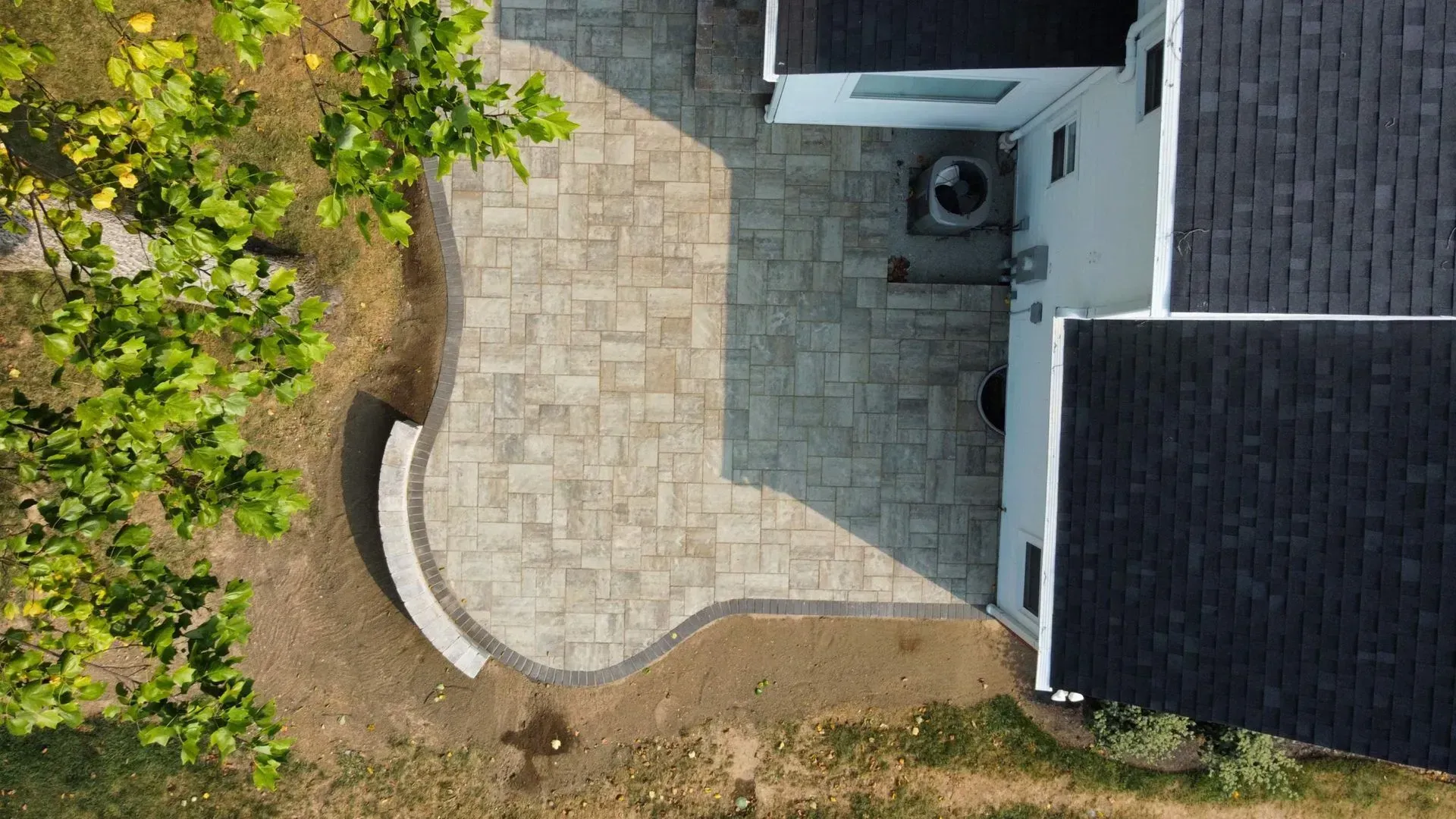 Overhead view of a curved stone patio beside a house and lawn, with an outdoor AC unit nearby