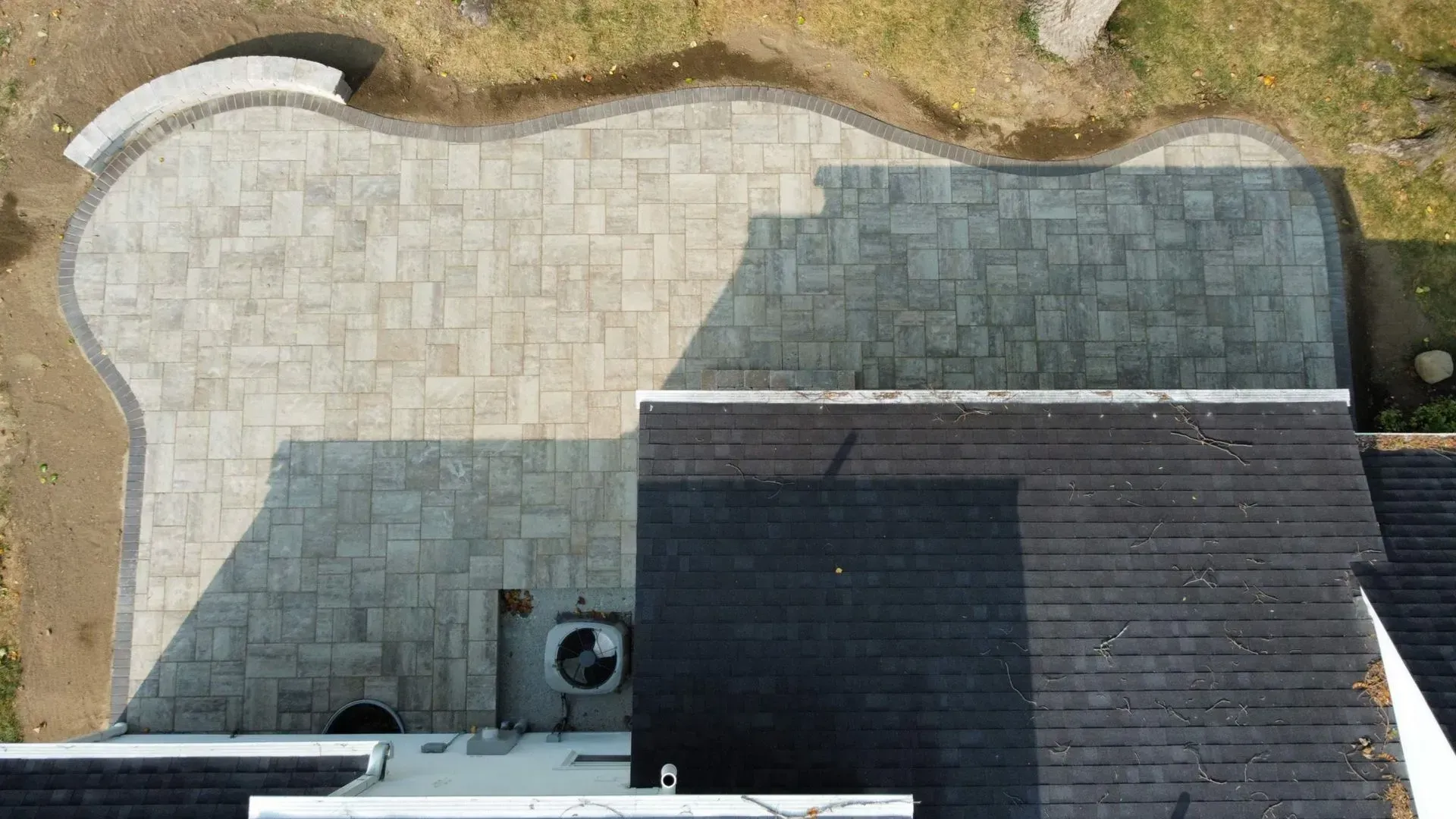 Aerial view of a stone patio beside a dark-roofed house and patchy lawn.