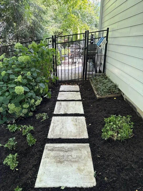 Narrow garden path with stepping stones leading to a black gate beside a house and greenery