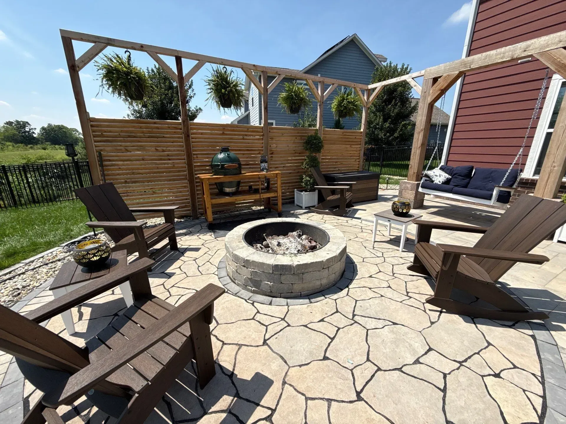Backyard patio with a stone fire pit, wooden chairs, grill, and pergola under a sunny sky