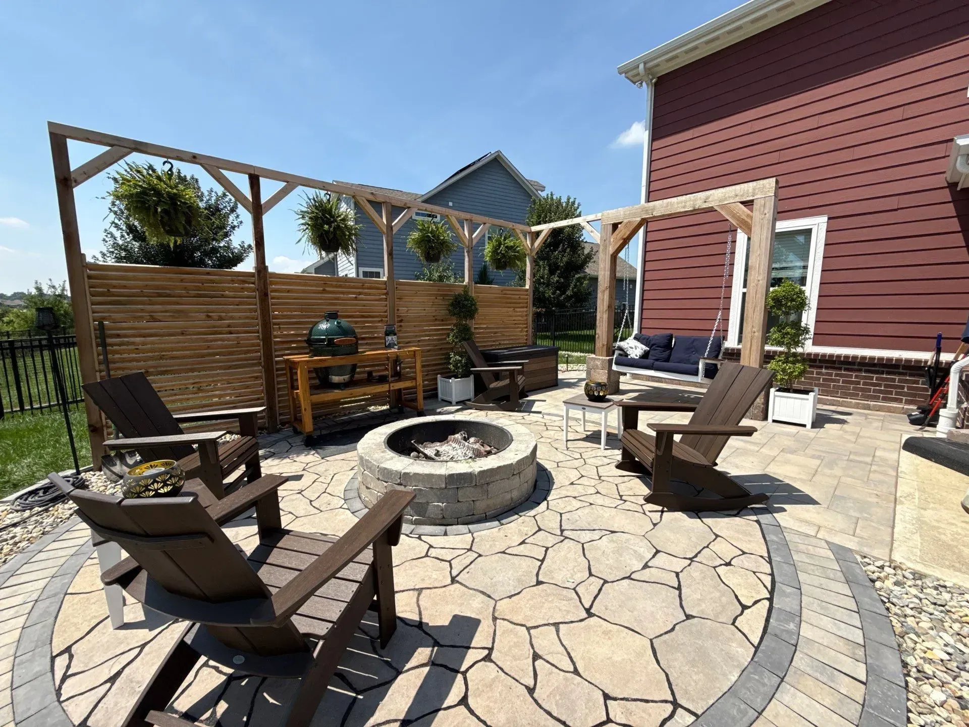 Backyard patio with stone fire pit, wooden chairs, and pergola seating beside a house