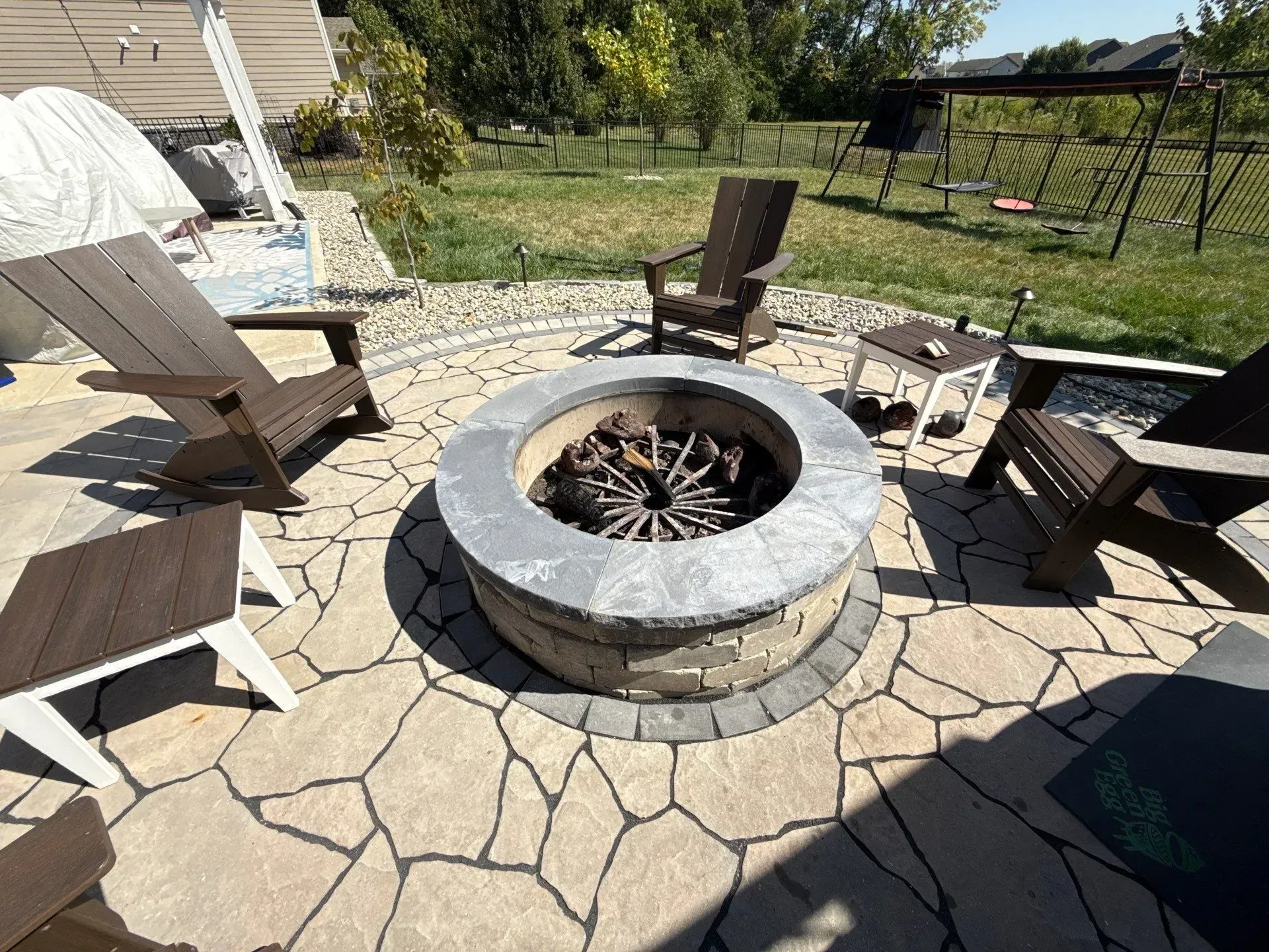 Stone fire pit surrounded by outdoor chairs on a patio with a grassy backyard beyond
