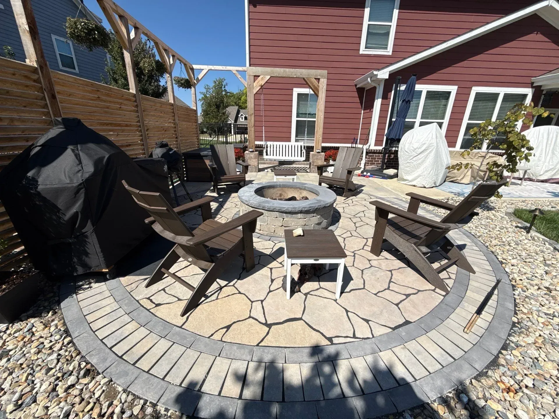 Backyard patio with circular stone fire pit, chairs, grill, and red house exterior
