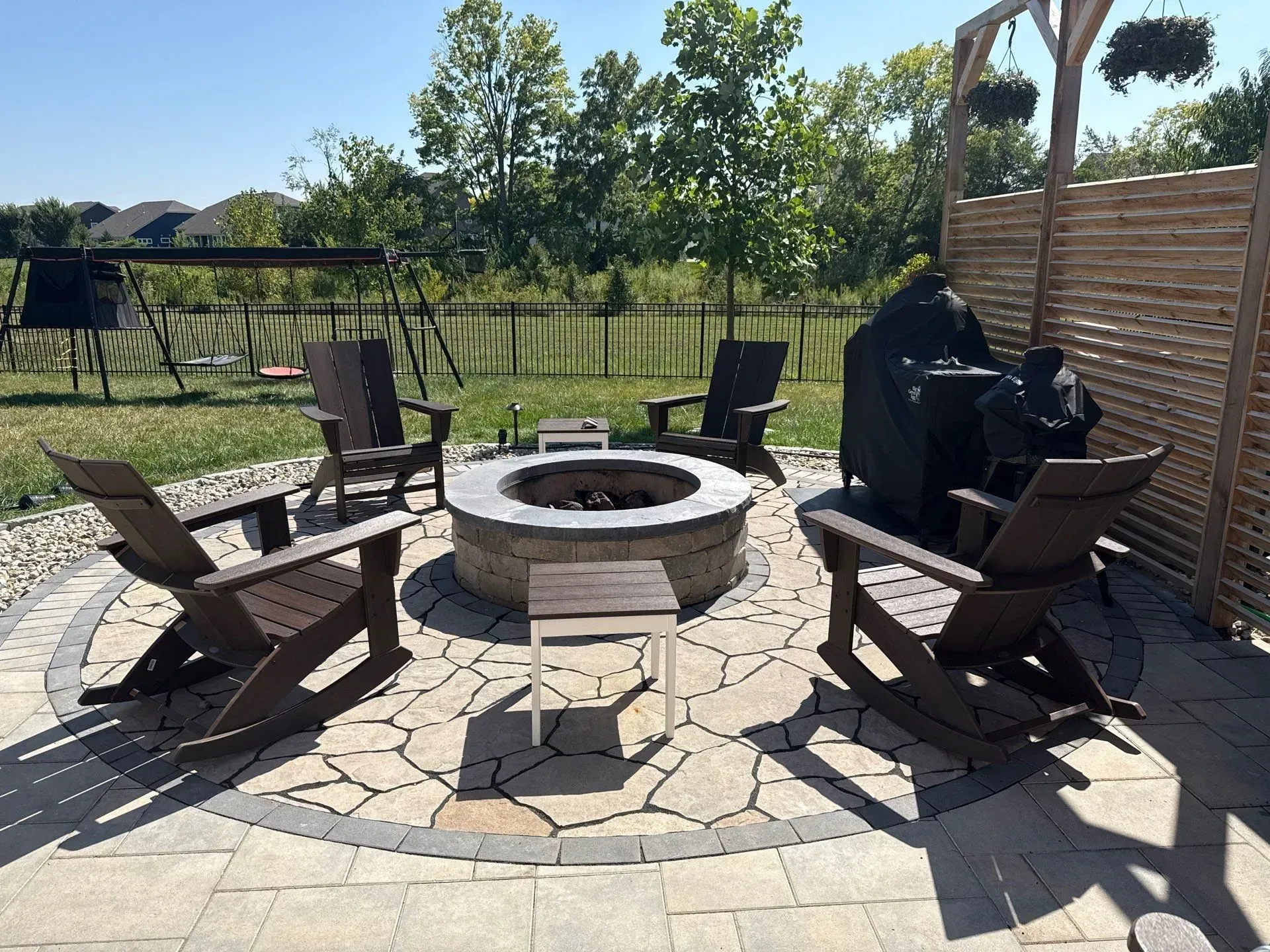 Patio fire pit seating area with brown chairs, stone pavers, and a grill in a sunny backyard