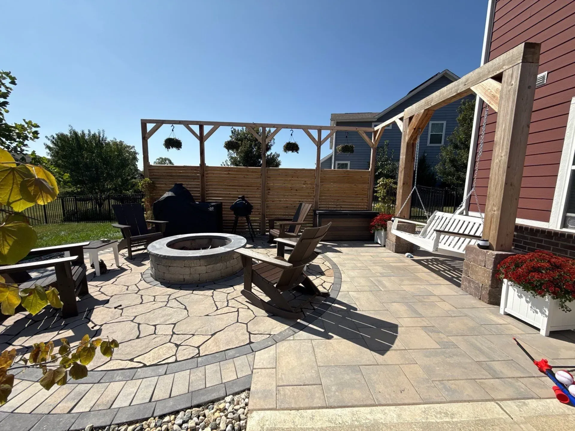 Backyard patio with fire pit, Adirondack chairs, potted flowers, and wooden privacy fence under blue sky