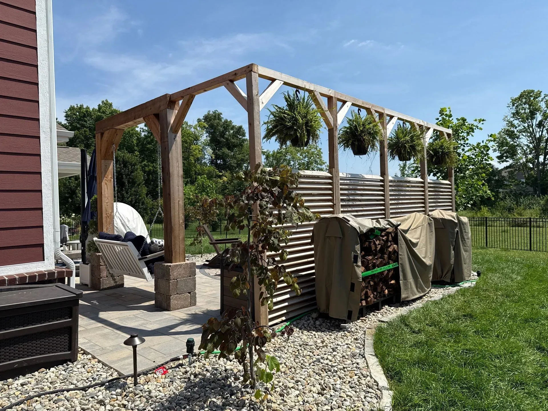 Backyard patio with wooden pergola, chairs, and metal privacy panels beside a house and lawn.
