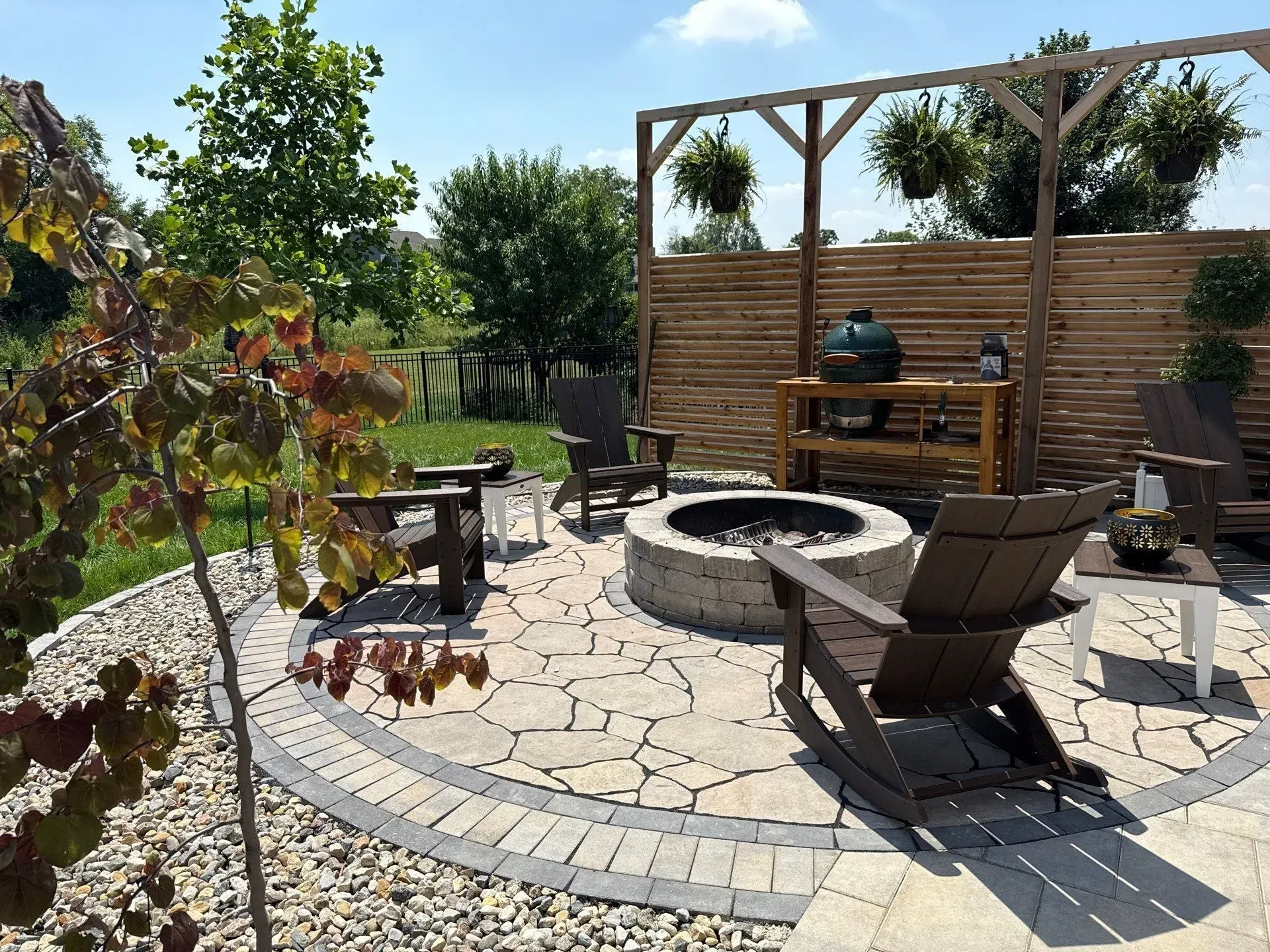 Stone patio with fire pit, lounge chairs, and a wooden privacy fence in a sunny backyard.