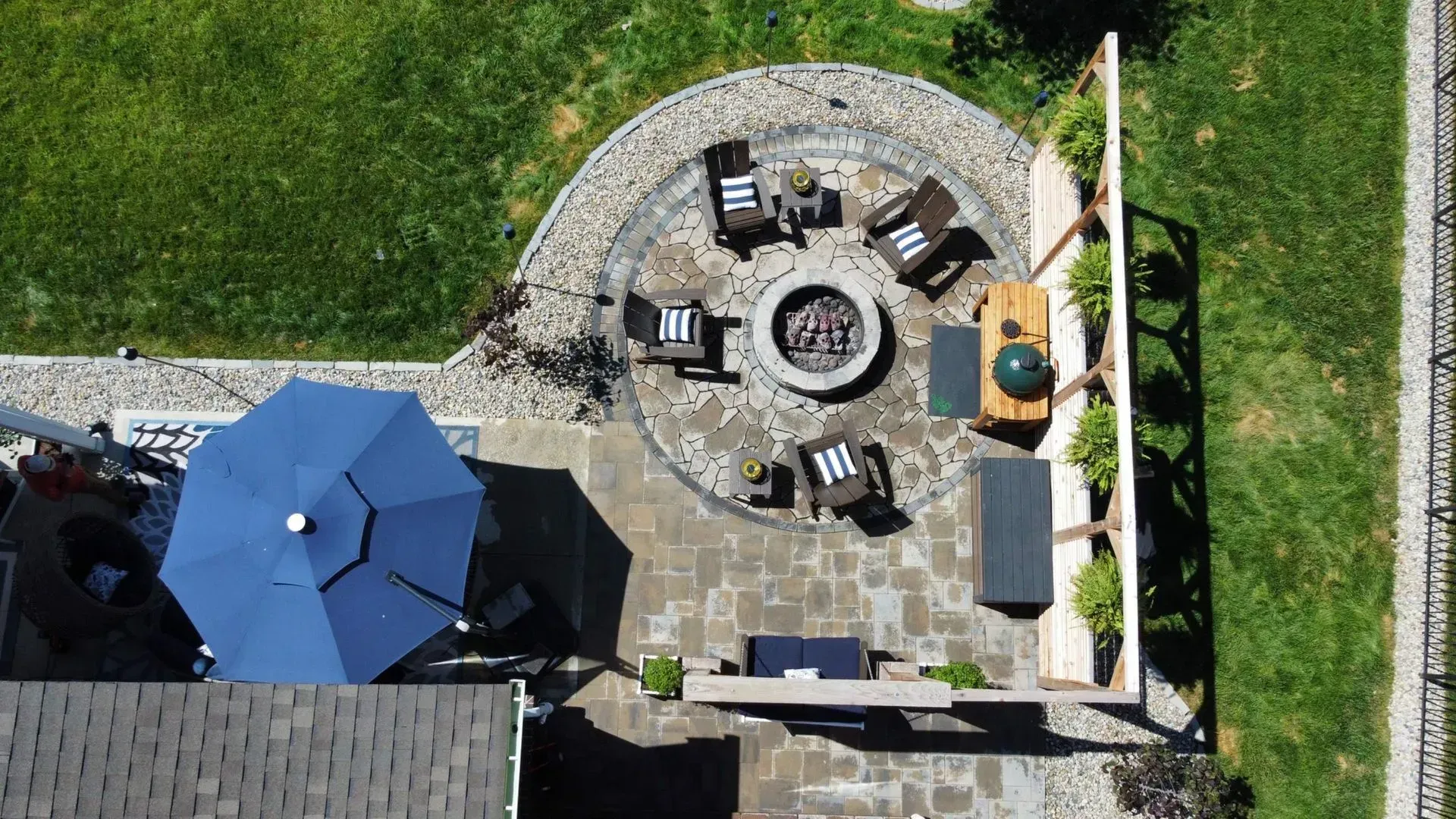 Aerial view of a backyard patio with blue umbrella, fire pit seating, and a grill beside a green lawn.