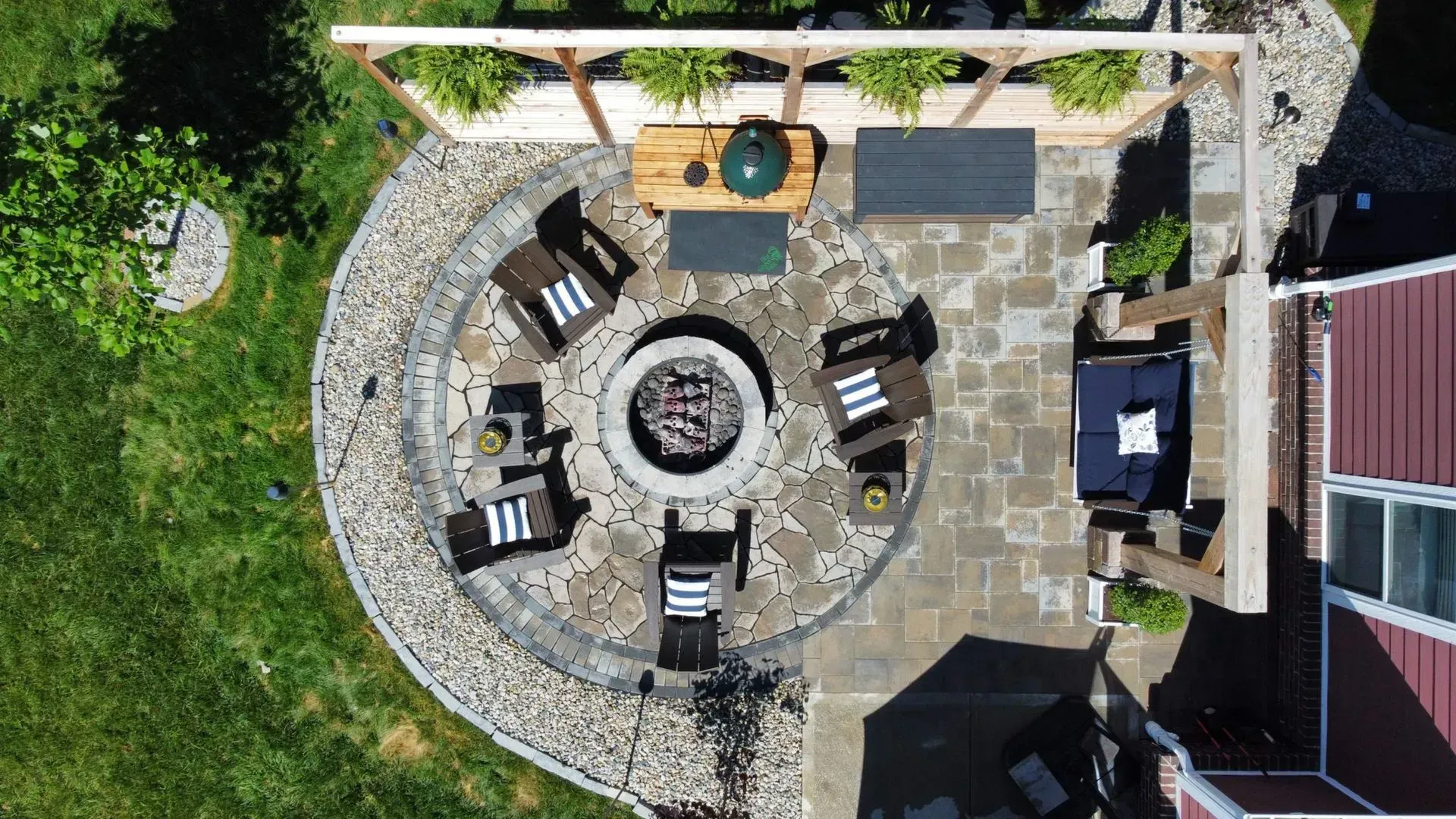 Overhead view of a stone patio with a fire pit, lounge chairs, and outdoor seating beside a house and lawn