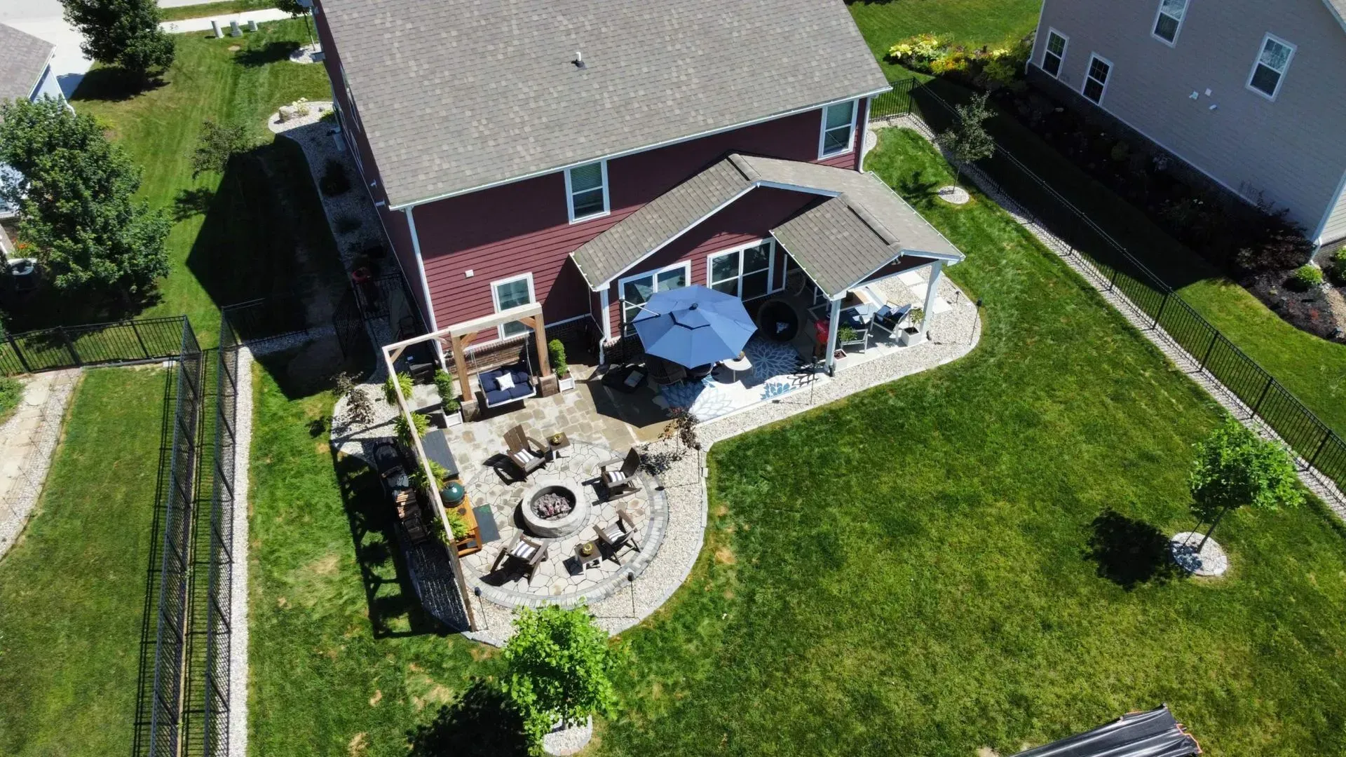 Aerial view of a backyard patio with a fire pit, blue umbrella, and red house beside a green lawn.
