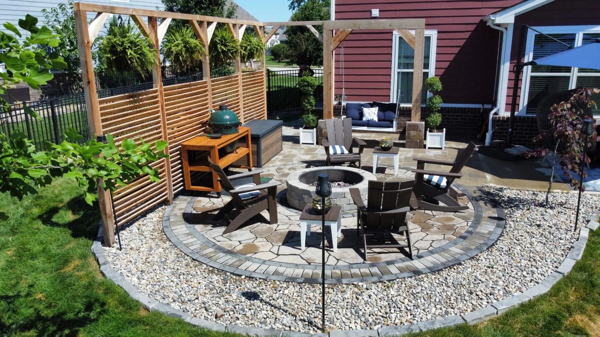 Backyard patio with circular stone fire pit, outdoor seating, and wooden fence under a pergola