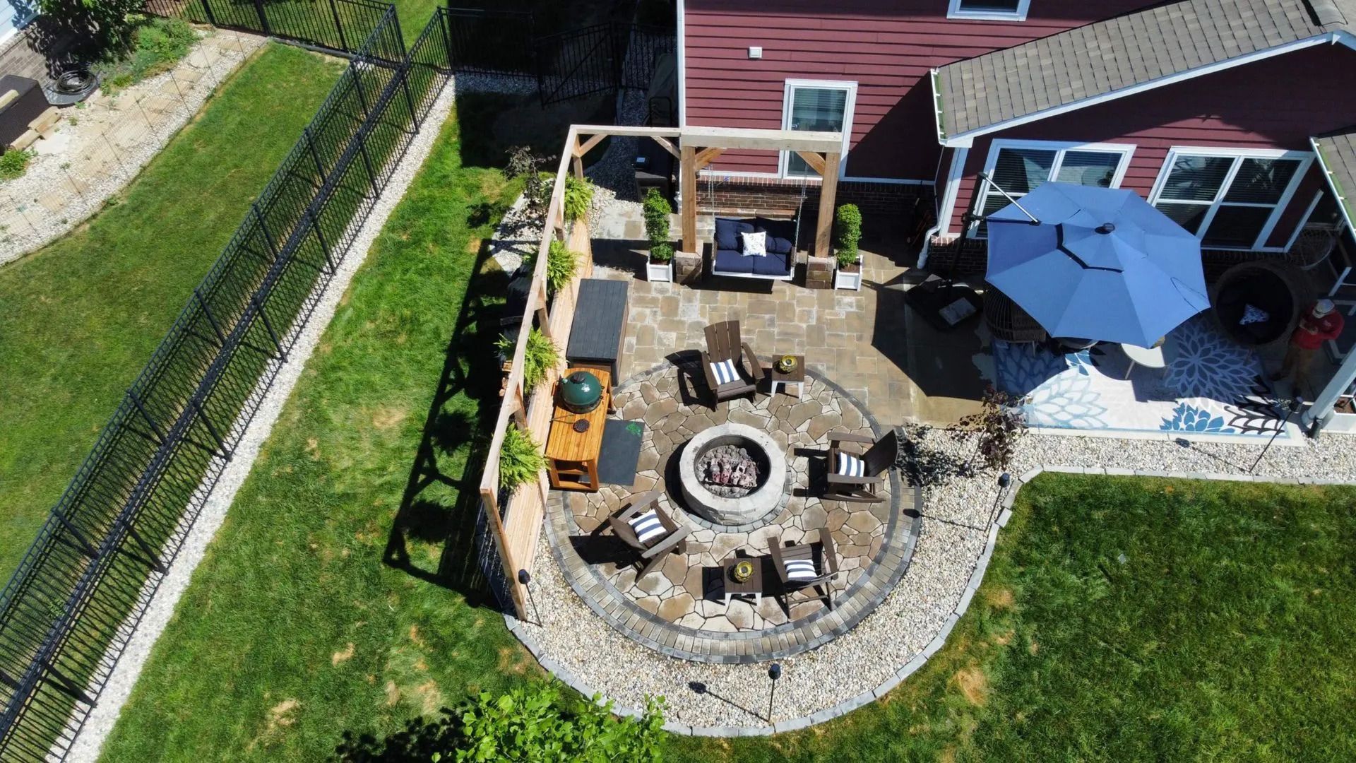 Backyard patio with circular stone fire pit, black chairs, wooden pergola, and surrounding greenery