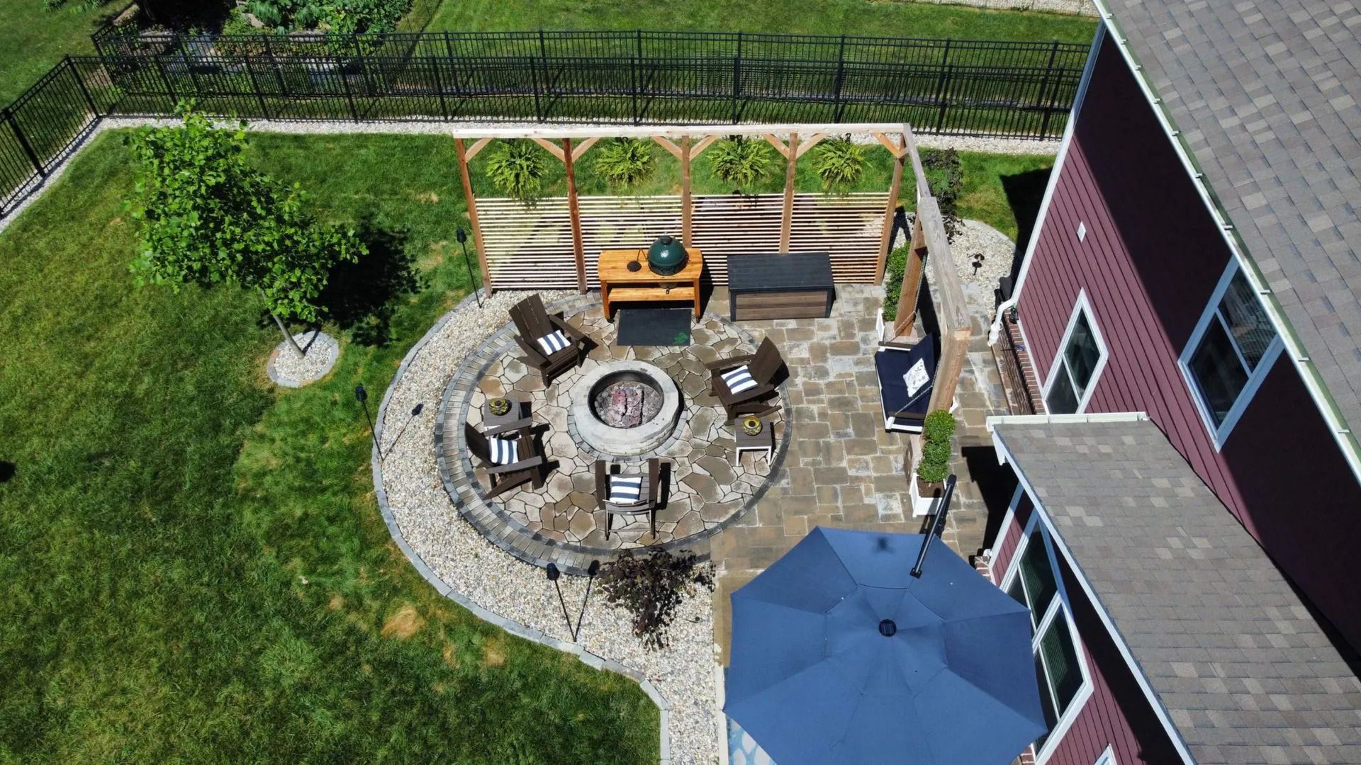 Backyard patio with a round fire pit, chairs, grill, and blue umbrella beside a house and lawn