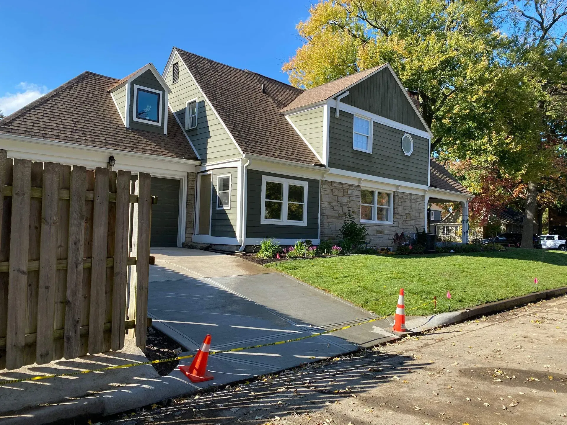 House with driveway under construction, orange cones by a wooden fence and green lawn on a sunny day