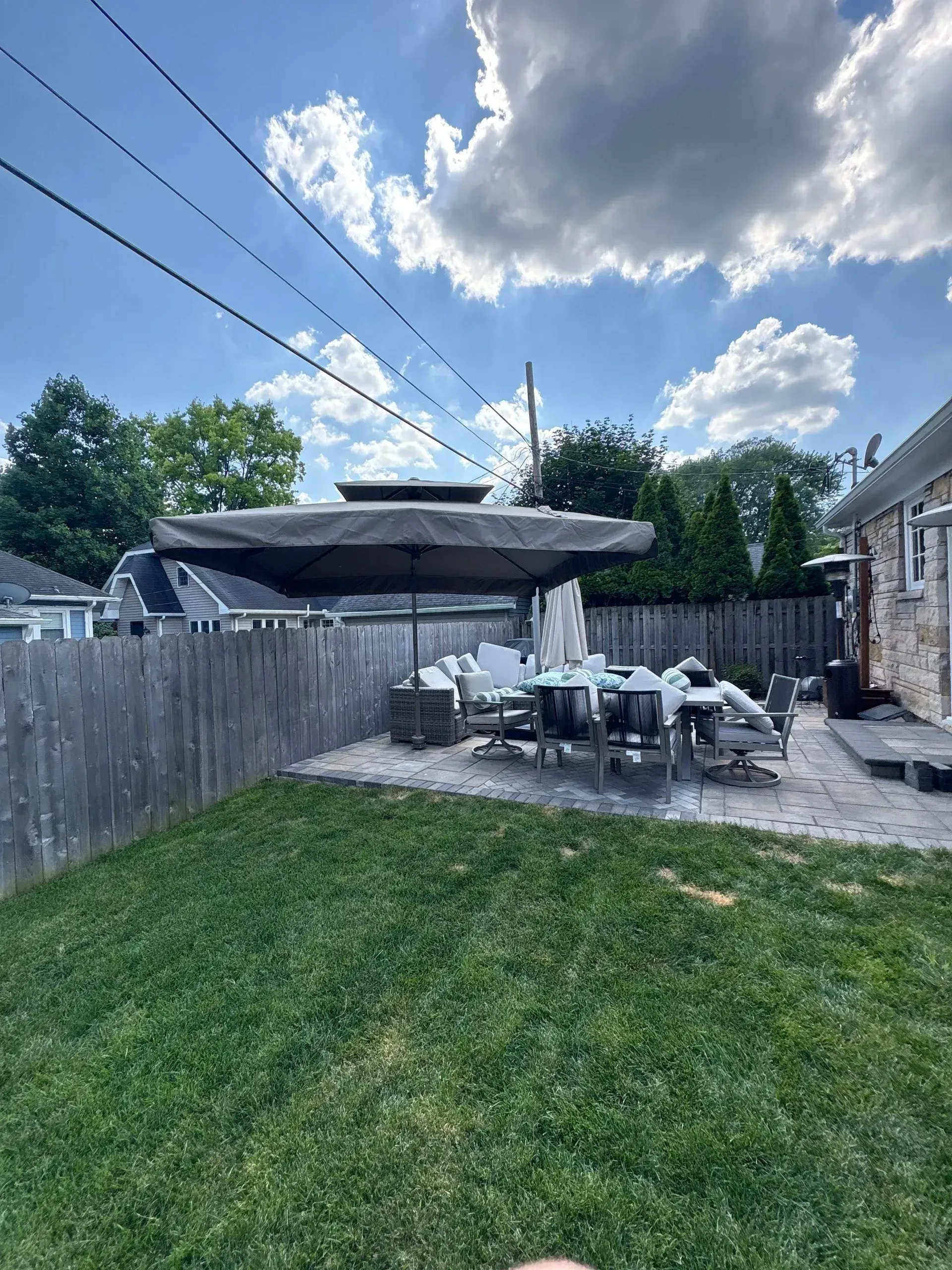 Backyard patio with white furniture, large umbrella, wooden fence, and green lawn under a blue sky.