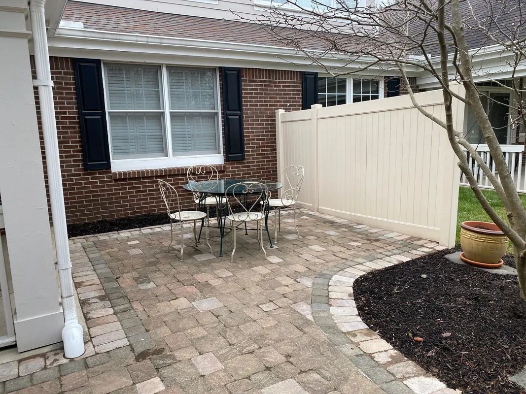 Brick patio with outdoor table and chairs beside a house, white fence, and potted plant.